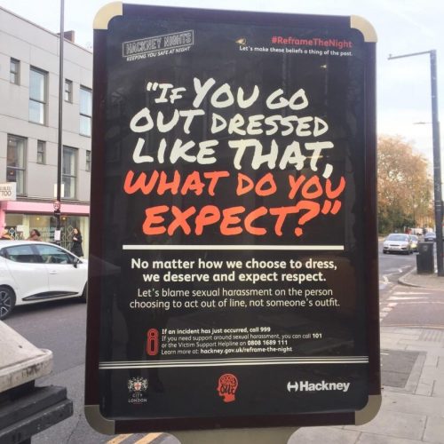 A bus stop near Dalston Junction station displaying a Reframe The Night poster. In the top-left corner is the Hackney Nights logo, with the slogan 'Keeping you safe at Night', while the top right has the #ReframeTheNight hashtag, with 'Let's make these beliefs a thing of the past' underneath. Large text in a handwriting font then reads, in quotation marks, 'If you go out dressed like that, what do you expect?'. Underneath, in a smaller sans-serif font, text reads 'No matter how we choose to dress, we deserve and expect respect. Let's blame sexual harrassment on the person choosing to act out of line, not someone's outfit.' At the bottom of the poster are the City of London, Good Night Out and Hackney Council logos.